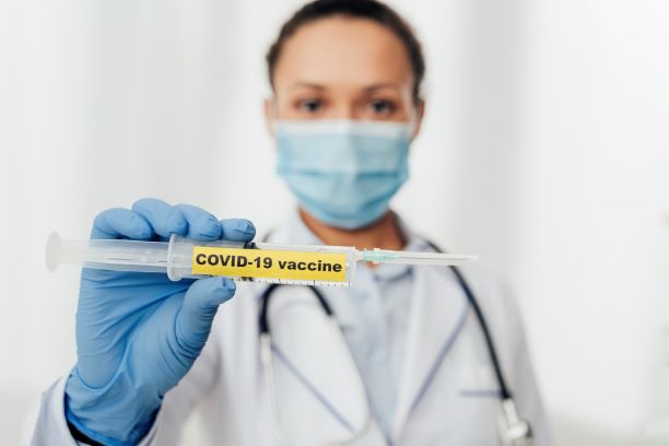 Kobieta w maseczce chirurgicznej na twarzy trzymająca w dłoni strzykawkę podpisaną Covid-19 vaccine.